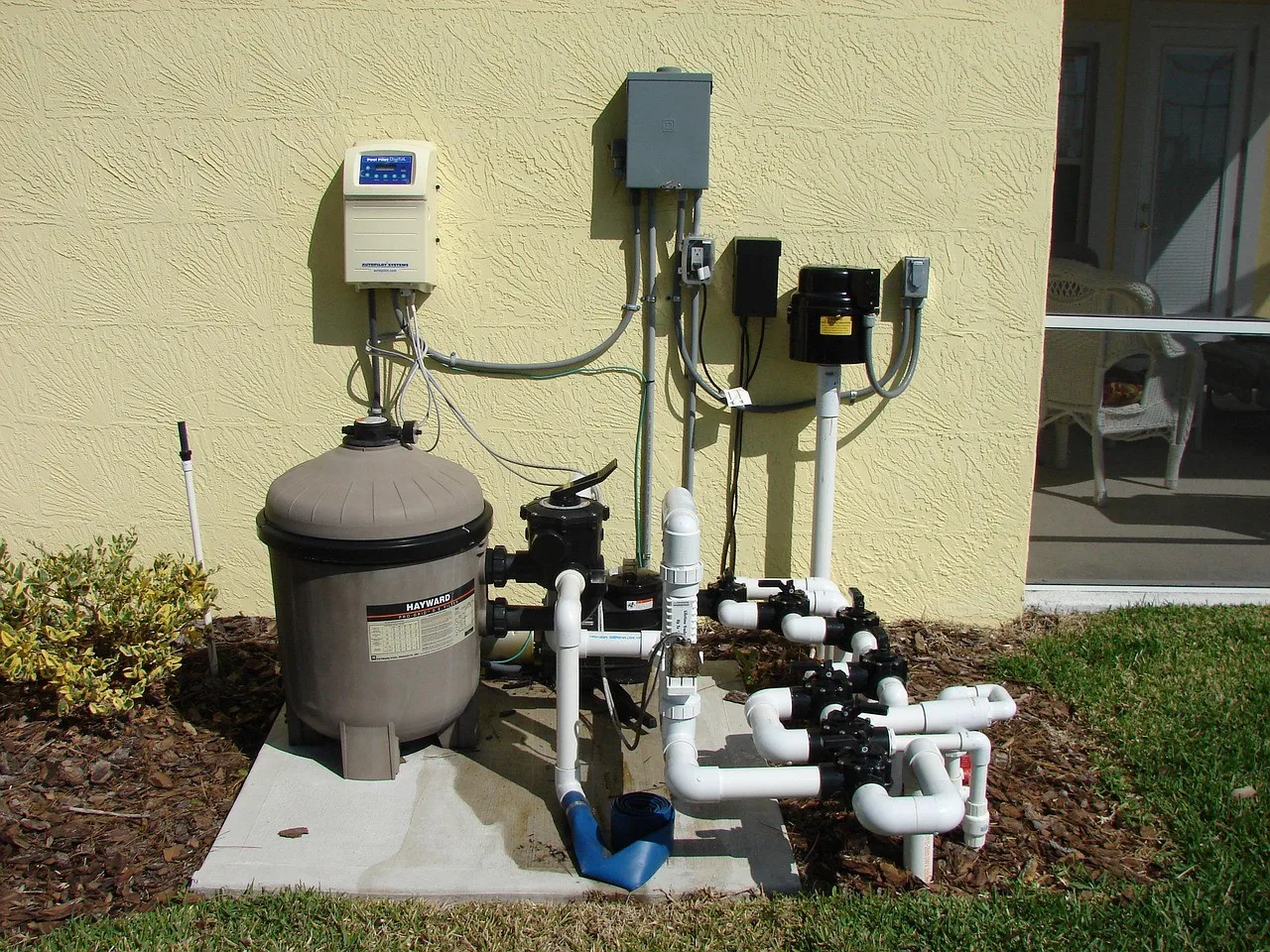 Outdoor pool pump and filtration system with multiple white PVC pipes, control boxes, and a round filter unit installed on a concrete pad next to a yellow stucco wall. For expert setup in MA, contact a trusted plumber Plymouth residents recommend.