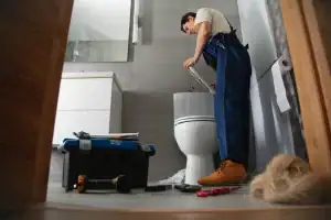 A plumber in blue overalls repairs a toilet in a modern Suffolk County, MA bathroom, with tools and a toolbox on the floor. A roll of toilet paper and a bundle of blonde hair are also visible.