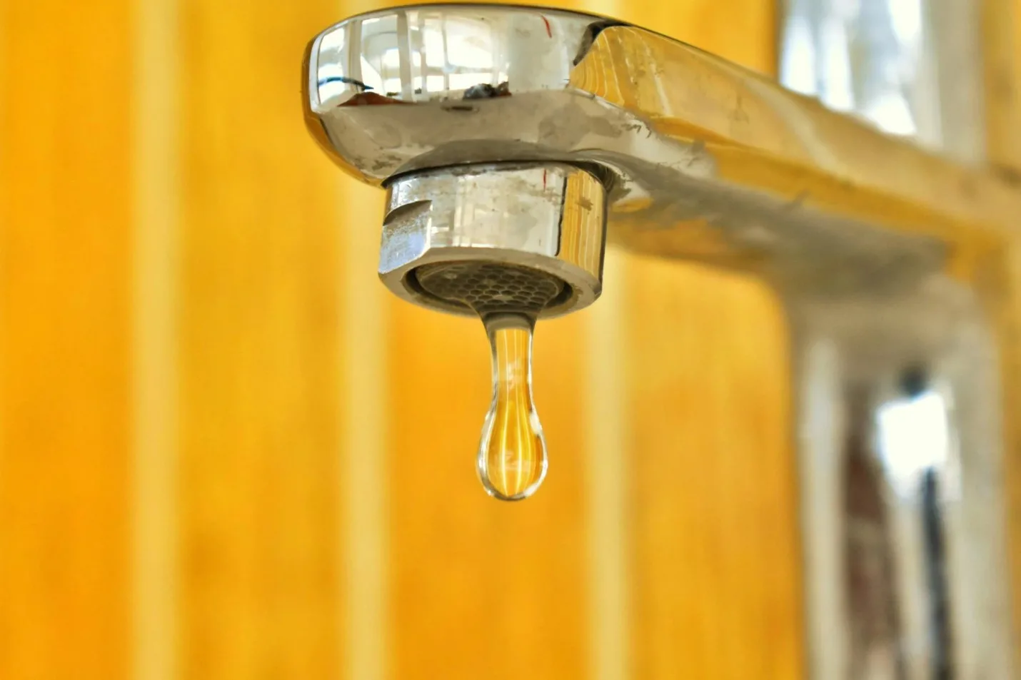 A close-up of a silver faucet with a single droplet of water hanging from its spout, set against a yellow and orange striped background—perfect for a plumber Plymouth or Bristol professional in MA seeking striking visuals.