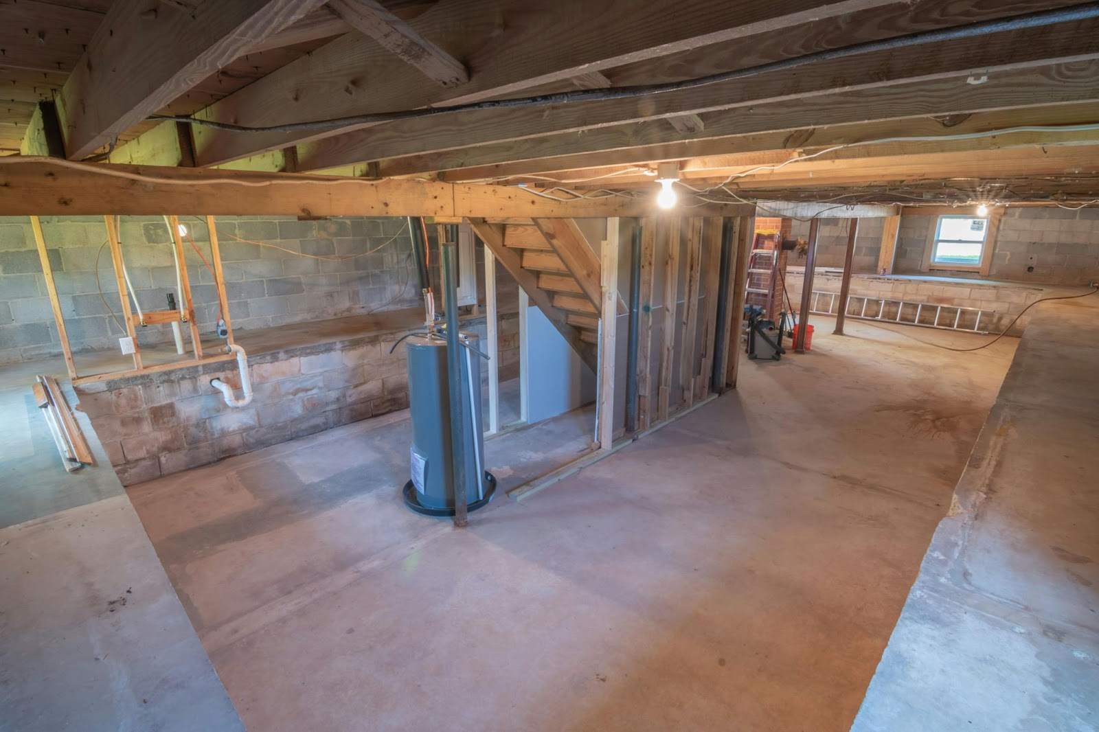Unfinished basement in Norfolk, MA, with exposed wooden beams, concrete floor, cinder block walls, open framing, a water heater, visible plumbing, and stairs leading up. A single light bulb provides illumination. Located near Suffolk County.