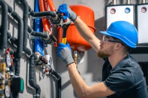 A worker in a blue hard hat, safety glasses, and gloves uses a wrench to adjust pipes connected to a complex plumbing or heating system on a wall in Middlesex, MA.