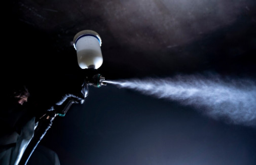 A close-up of a person using a spray gun to apply a mist of paint or coating, with the spray visibly dispersing in the air against a dark background—captured at a workshop in Bristol.