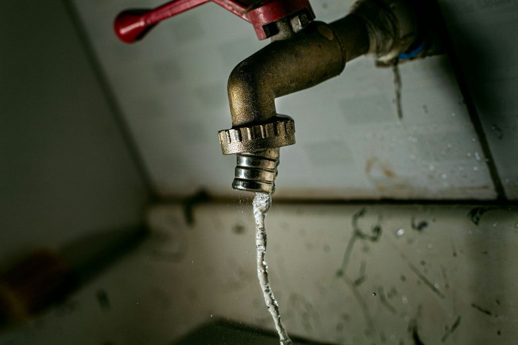 A close-up of a metal faucet with a red handle, partially open, letting out a thin stream of water into a dirty sink in Norfolk and Suffolk County.