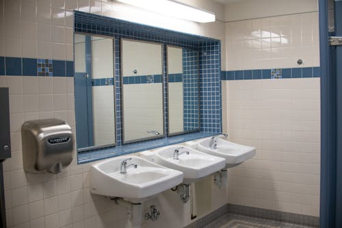 A clean public restroom in Middlesex features three white sinks, a large mirror above them, a stainless steel hand dryer on the wall, and blue and white tiled walls.