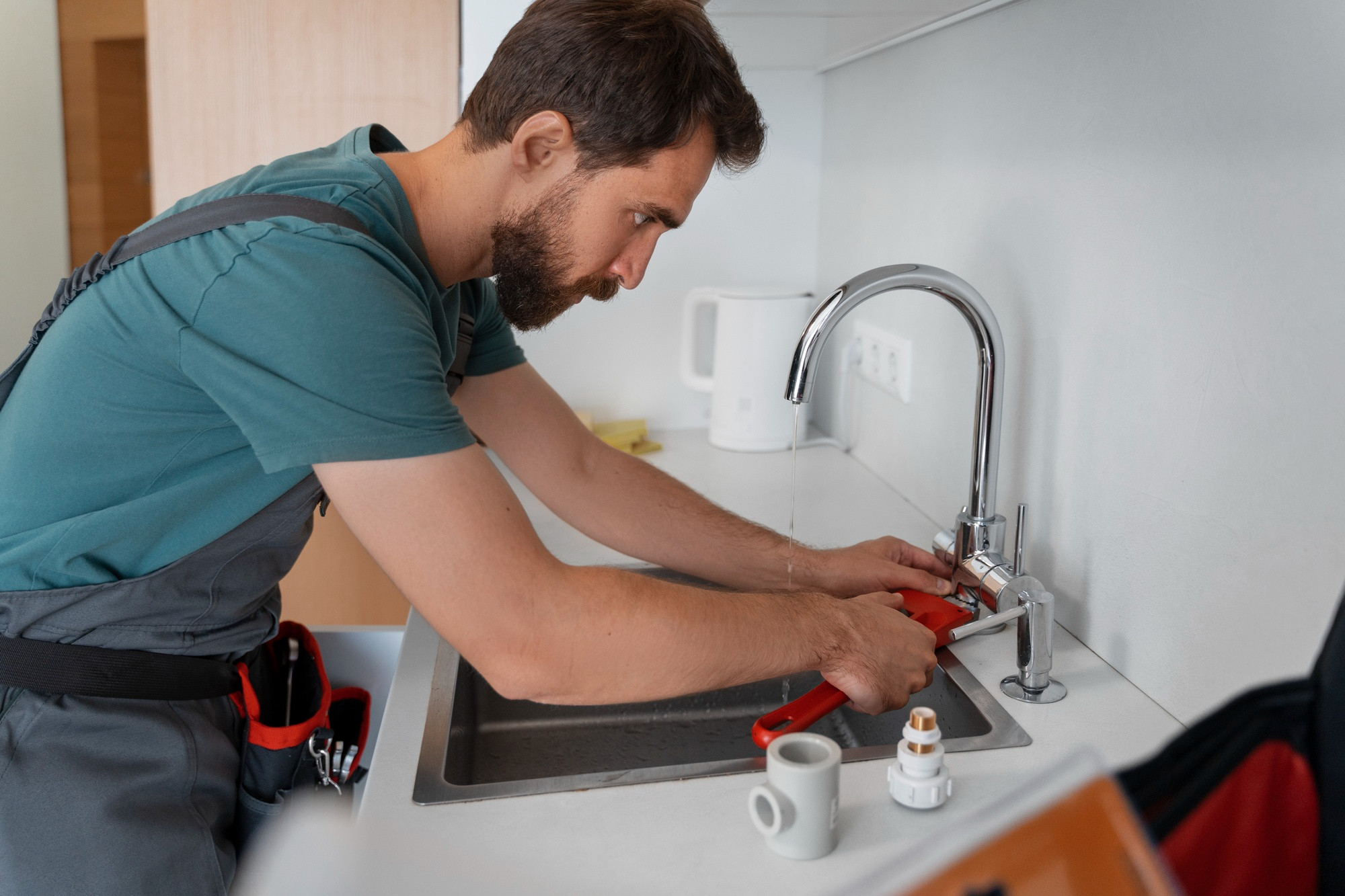 A plumber from Norfolk with a beard is using a red wrench to fix a kitchen faucet. He wears a teal shirt and overalls, with tools in his pocket. A white kettle and cup sit on the countertop beside him.