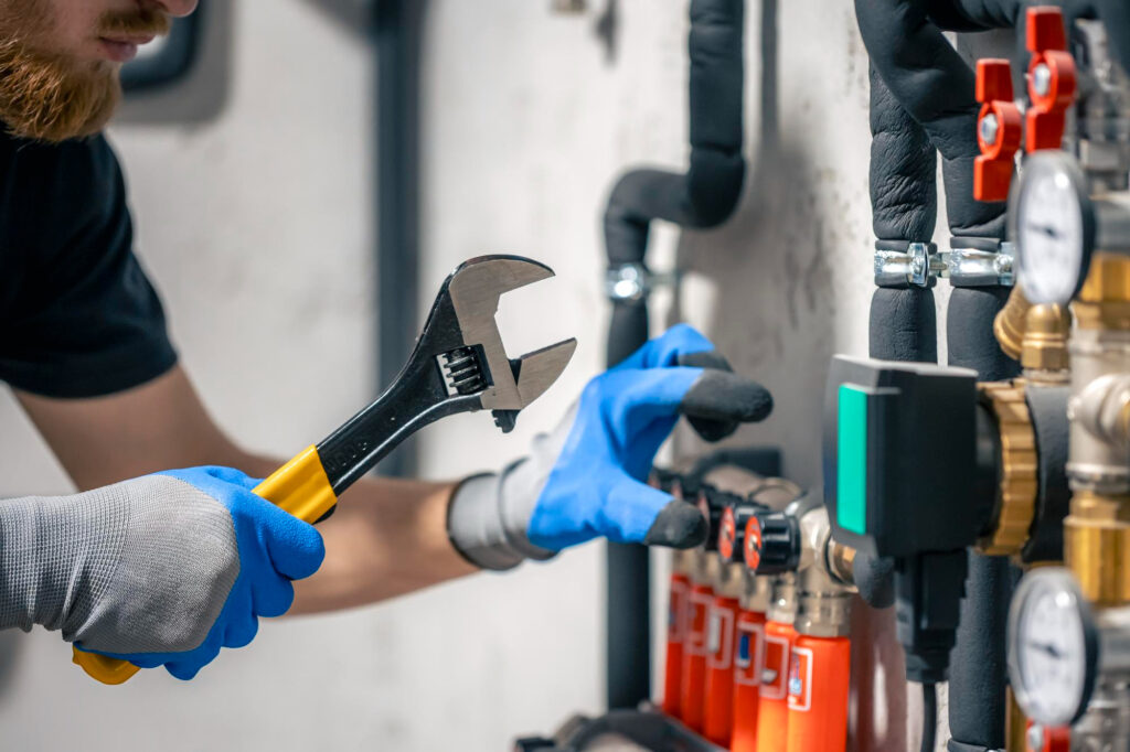 A plumber Plymouth, wearing blue gloves, uses a wrench to adjust pipes and valves on a heating system, with gauges and insulated pipes visible in the background.