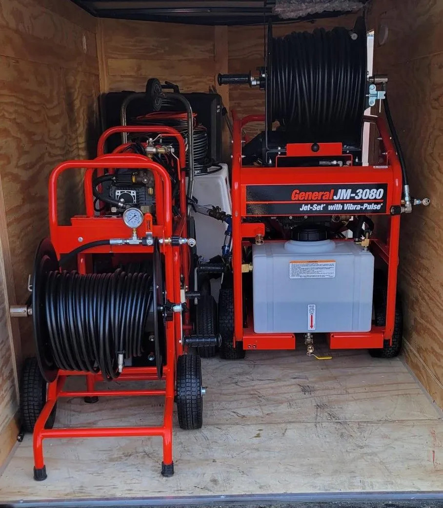 Two red and black industrial hose reel machines are stored inside a wooden trailer. The machine on the right is labeled "General JM-3080." Both machines have hoses wound on reels, ready for any plumber Plymouth or Suffolk County, MA might need.