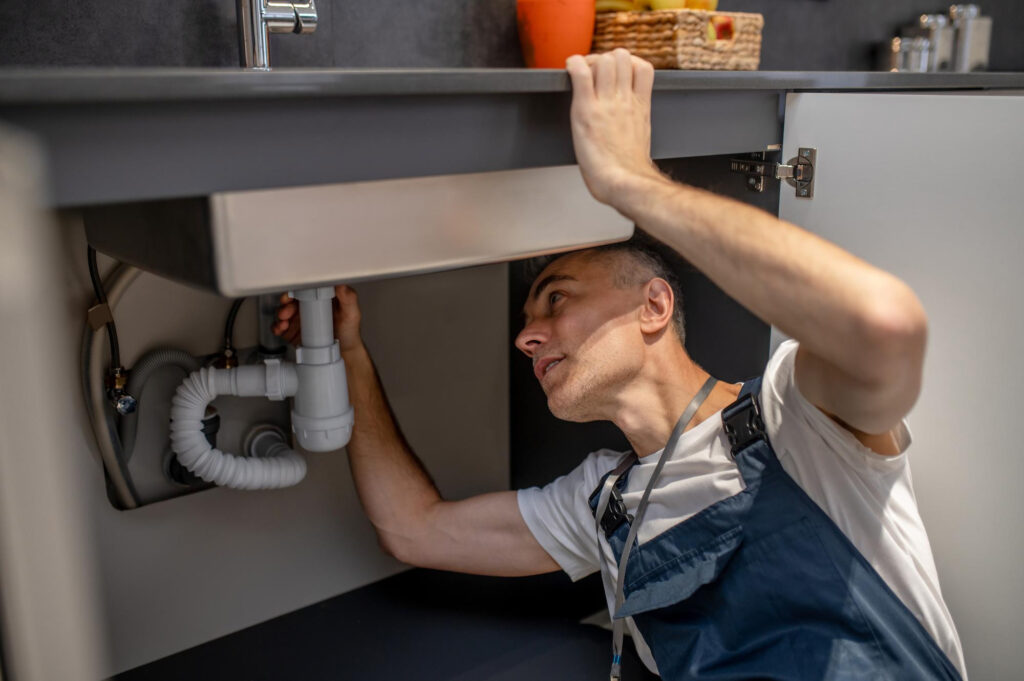 A man wearing overalls is inspecting the plumbing under a kitchen sink in a home located in Bristol County, MA, with cabinet doors open and various pipes visible.