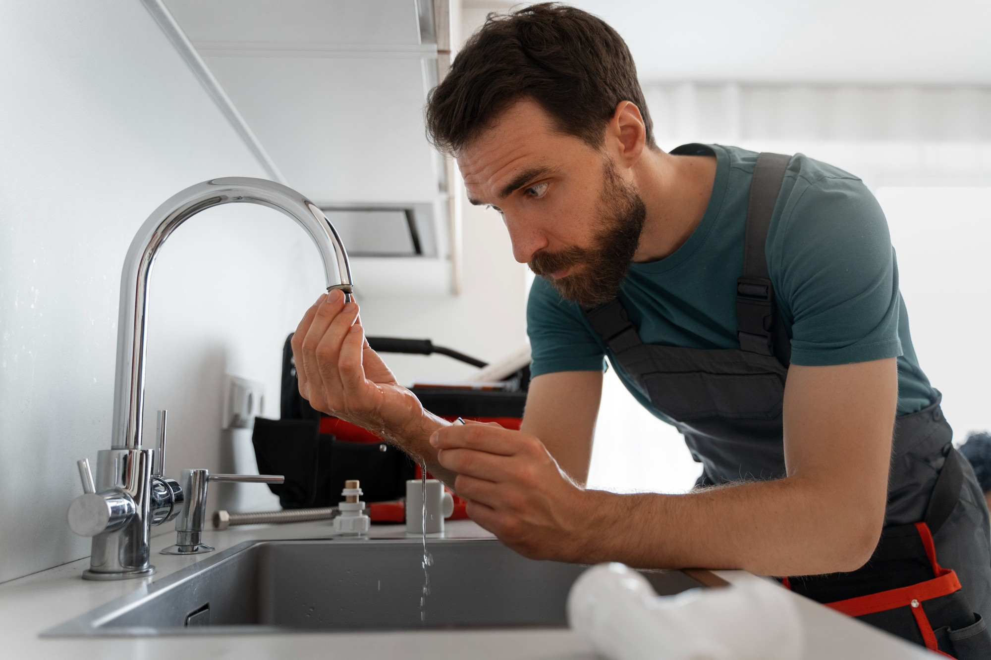 A plumber in overalls examines a kitchen faucet in Middlesex, MA, checking for leaks while water drips from the tap. He is focused, holding a small part, with tools and a tool bag visible in the background.