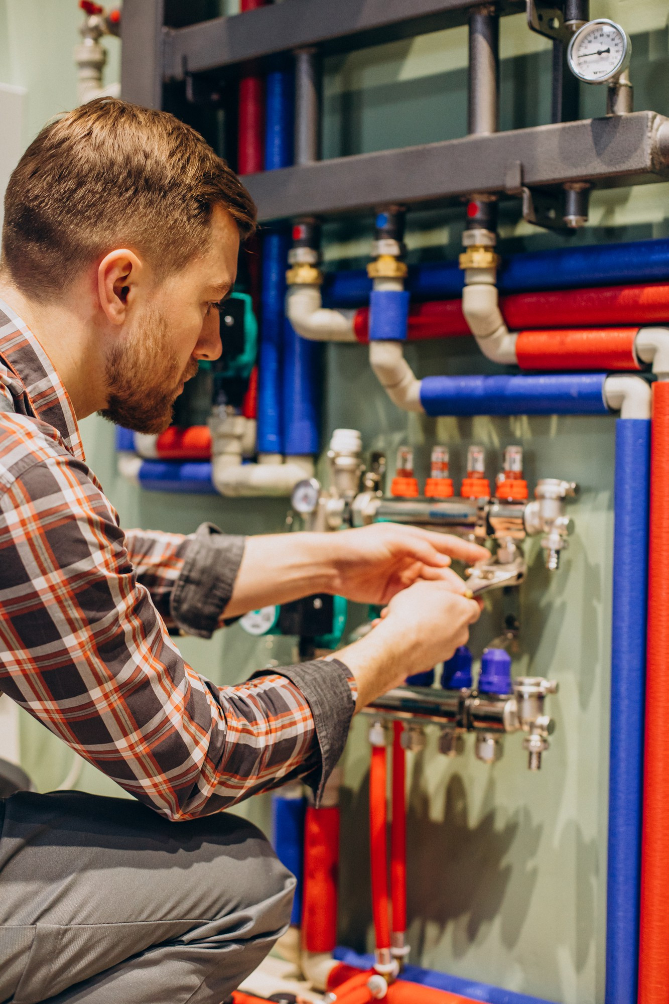 A plumber from Plymouth in a plaid shirt kneels while adjusting valves on a panel connected to red and blue pipes, likely working on a plumbing or heating system. A pressure gauge is visible at the top right.
