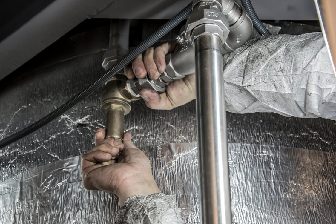 A person in protective clothing is using both hands to tighten or install metal plumbing pipes and fittings beneath a reflective insulated surface, completing important work in Middlesex.