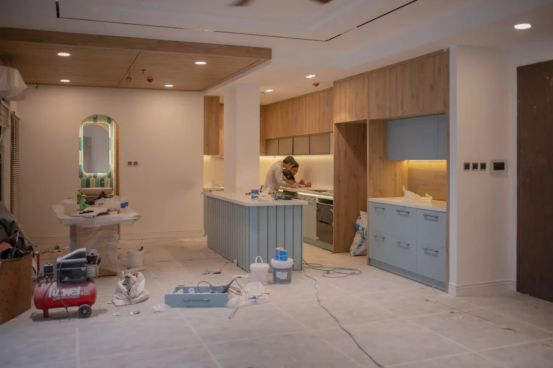 A man works in a partially finished kitchen in MA, with light wood cabinets, blue drawers, and construction tools scattered across the floor—capturing home renovation in progress common to Middlesex and Suffolk County projects.