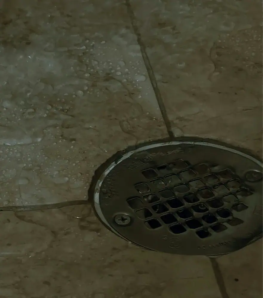 A metal shower drain set into brownish tile flooring, with water droplets and wet areas visible on the tiles around the drain—typical of bathrooms in homes across Middlesex and Suffolk County.