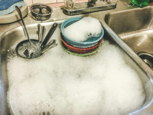 A kitchen sink filled with soapy water, foam, and a stack of colorful bowls—just another busy day in Middlesex. Several spoons and utensils are submerged in the soapy water, ready to be washed.