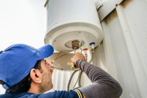 A plumber from Plymouth wearing a blue cap repairs a water heater mounted on an exterior wall, using a screwdriver to work on exposed wires and components underneath the tank.