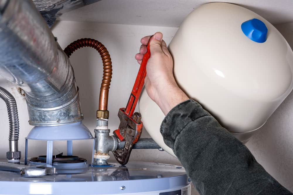 A person in Norfolk uses a red pipe wrench to tighten or loosen a fitting on a water heater system, with metal and copper pipes visible and an expansion tank attached.