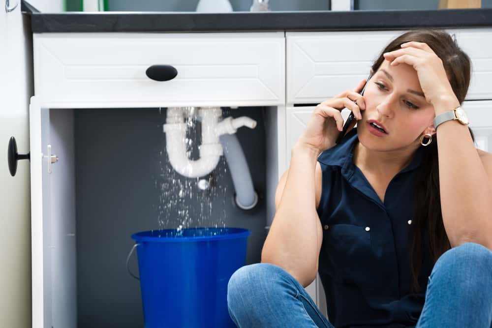 A woman sits on the floor by a kitchen cabinet in Suffolk County, MA, looking stressed while talking on the phone to a plumber. Water leaks from a pipe under the sink into a blue bucket.
