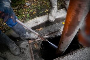 Two people in work boots and gloves use a hose to clean or clear a storm drain on a concrete street in Middlesex, MA, with leaves and debris visible nearby—an essential task for any skilled plumber.