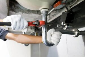 A person wearing white gloves uses a red pipe wrench to work on plumbing maintenance under a sink, just like a professional in Bristol, MA or Suffolk County.