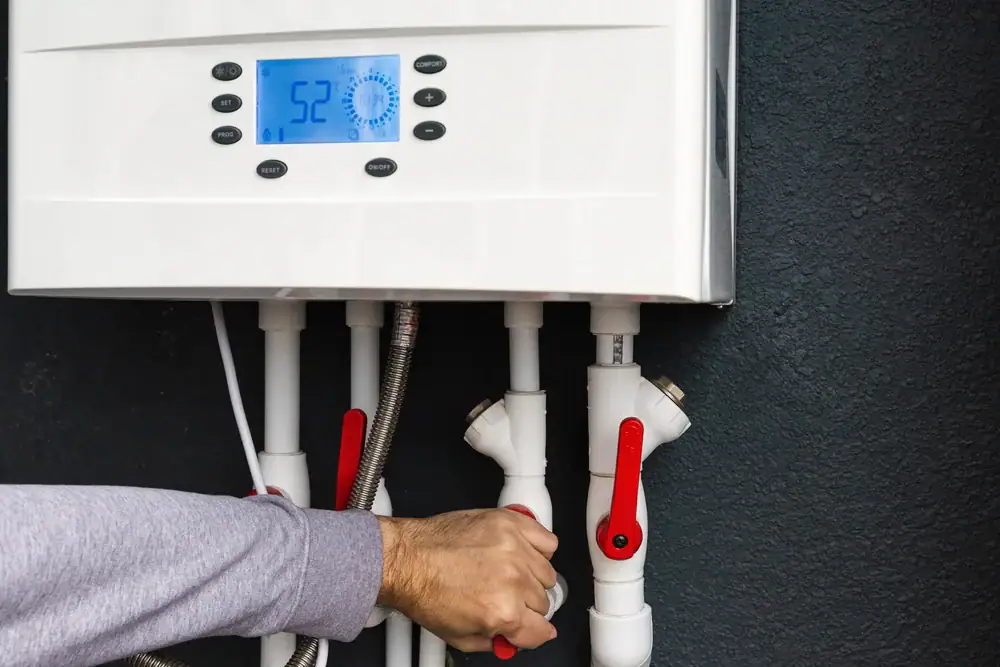A person adjusts a valve on a white wall-mounted water heater with pipes and a digital display showing the temperature as 52°C.