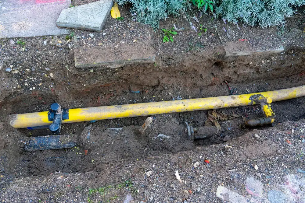 A yellow gas pipeline is partially exposed in a trench, surrounded by dirt and soil. Various fittings and valves are attached to the pipe, indicating ongoing maintenance or installation work.