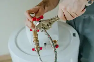 A person uses a wrench to tighten a pipe connection on top of a white water heater, focusing on plumbing installation or repair.