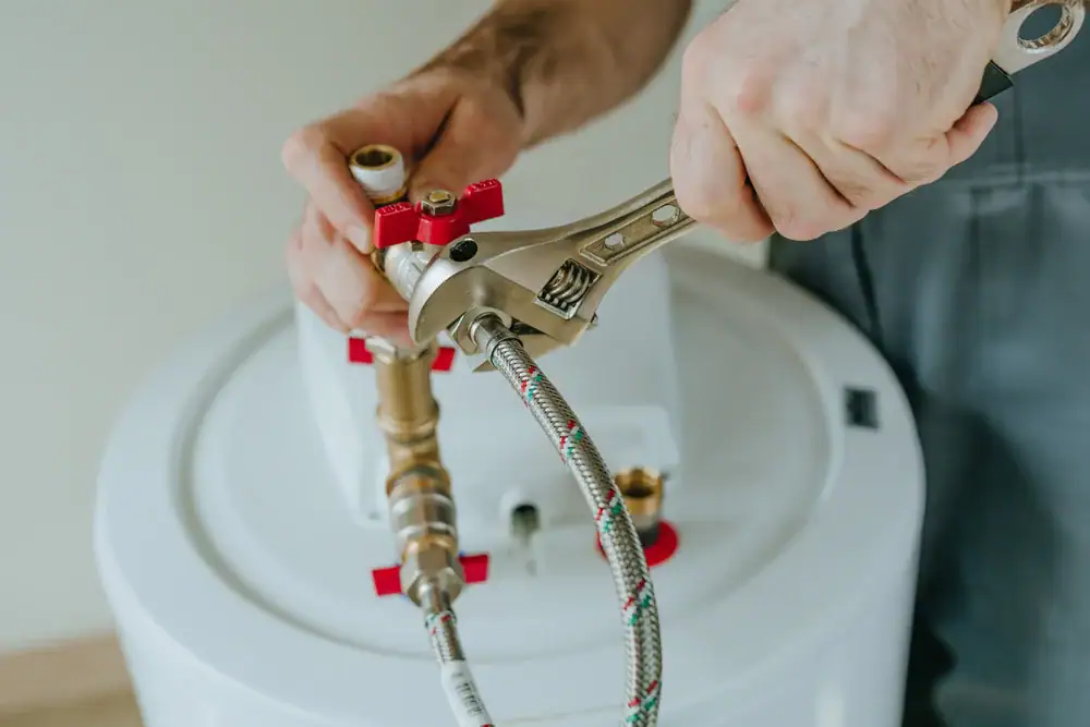 A person uses a wrench to tighten a pipe connection on top of a white water heater, focusing on plumbing installation or repair.