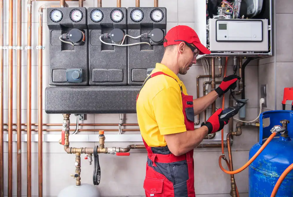 A technician in a yellow shirt, red cap, and red gloves uses a handheld device to inspect pipes and gauges on a heating system, surrounded by copper pipes and equipment in a utility room.