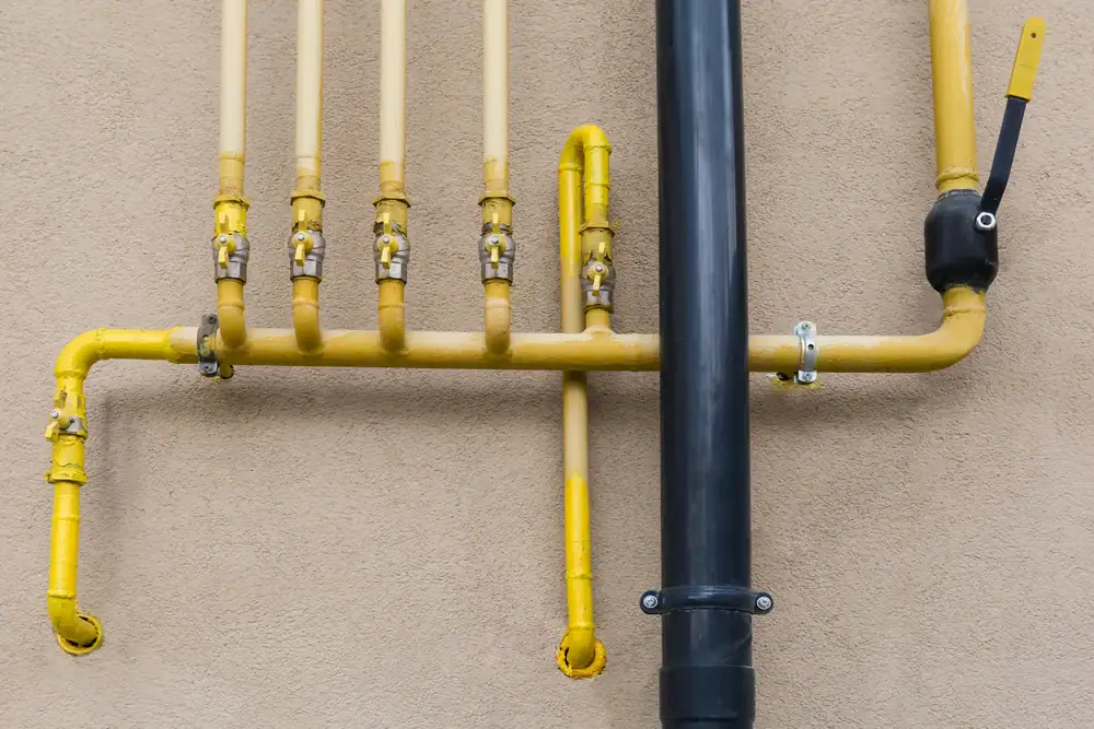 Yellow gas pipes with several valves run horizontally and vertically along a beige wall, intersected by a large black vertical pipe on the right side. A lever is attached to the black pipe.